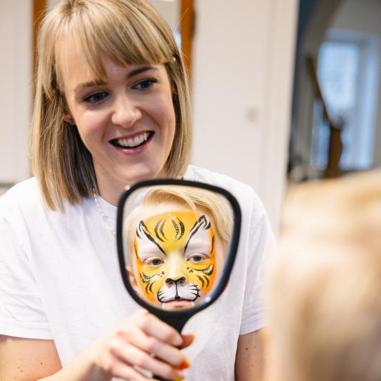 Kvinde holder spejl hvor dreng med ansigtsmalet tiger spejler sig