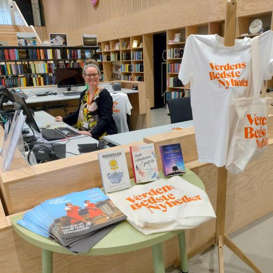 Billedet viser en udstilling med Verdens bedste nyheders avis, muleposer og t-shirts. Der ses også en bibliotekar bag skranken.