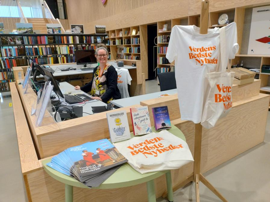 Billedet viser en udstilling med Verdens bedste nyheders avis, muleposer og t-shirts. Der ses også en bibliotekar bag skranken.