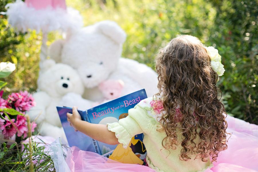 Billedet viser en pige, der læser en billedbog for sin bamse, mens de sidder ude i solen på græs.